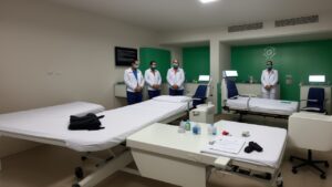 Profissional de saúde de jaleco conversa com paciente em sala de atendimento, representando a ética na saúde com livros de bioética ao fundo.