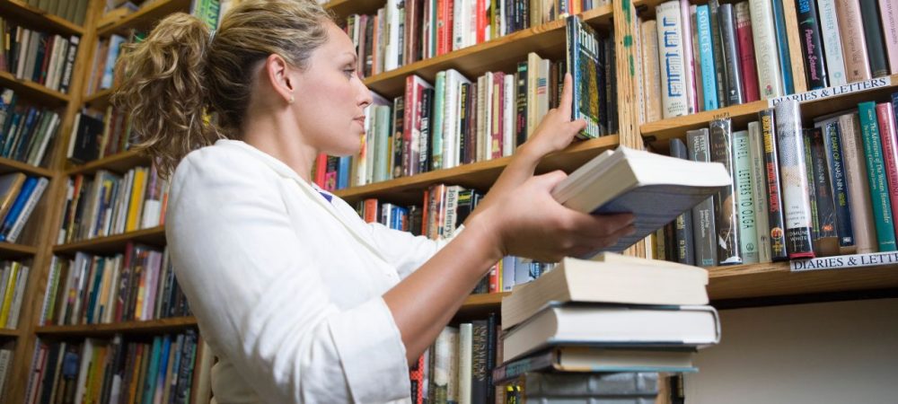 Pessoa organizando livros em biblioteca, ilustrando as atividades do auxiliar de biblioteca.
